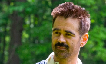 Jake portant une veste bleue, une chemise blanche et une écharpe blanche. Il regarde vers la gauche et a une moustache ainsi qu'une barbe.L’arrière plan est constité d'arbres flous.