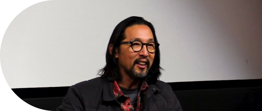 Kogonada portant des lunettes, une veste noire, une chemise à carreau rouge et un t-hirt gris. Il a les cheveux mi-longs et détaché ainsi qu'une moustache et une barbe. Derrière lui se trouve un mur blanc et noir.