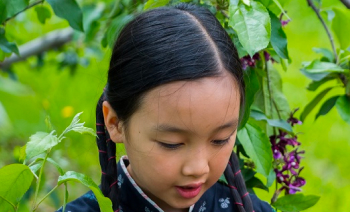 Mika au mileu de branche d'arbres. Elle regarde vers le bas et a deux tresses. Elle porte un vêtement chinois bleu marine avec des fleurs.
