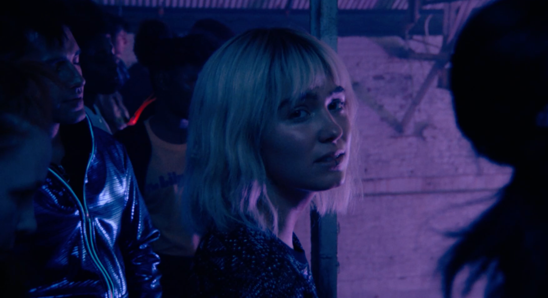 Ada se trouve dans un environement à la lumière mauve. D'autres personnes sont présentes autour d'elle. 