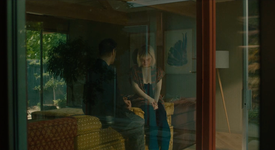 Jake et Ada discutent, assis sur le dossier d'un canapé. Nous les voyons à travers une baie vitrée. Jake est sur la gauche et nous tourne le dos. Ada est située sur la droite et nous fait face.