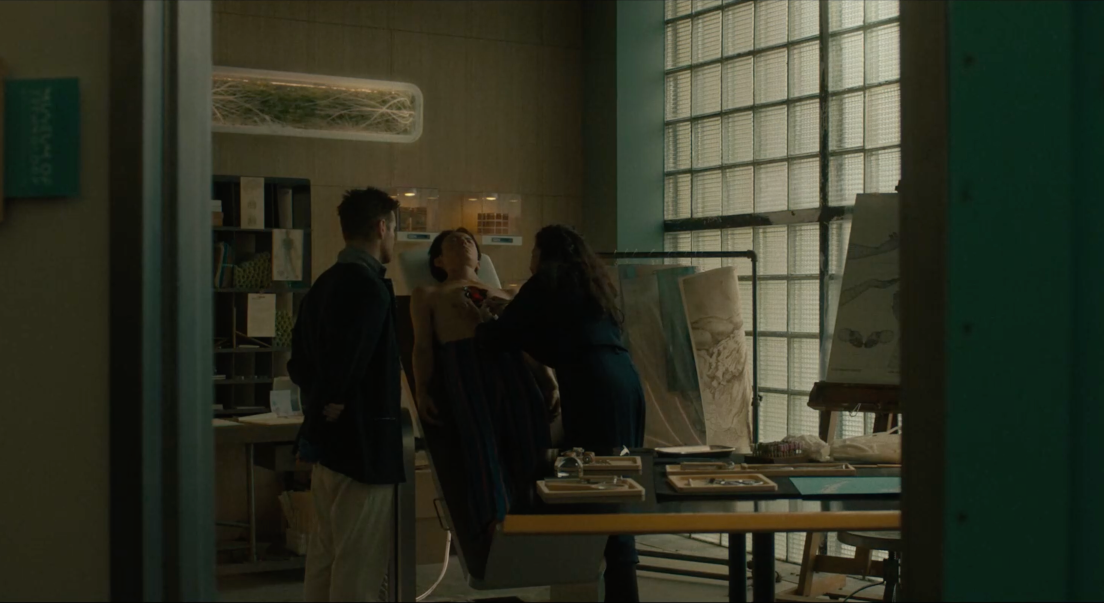 Cleo examine le corps de Yang, déposé sur une table verticale. Jake se trouve sur le côté et l'observe. Ils se trouvent dans un laboratoire scientifique.