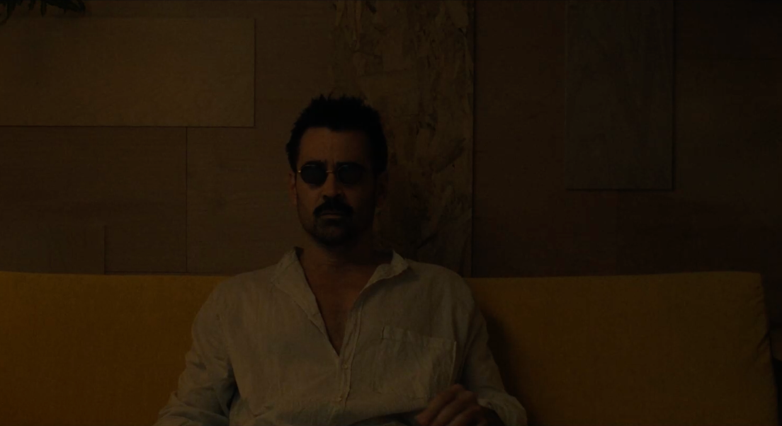 Jake est assis sur un fauteuil jaune, portant des lunettes de soleil et une chemise blanche. La pièce est plongée dans la pénombre.