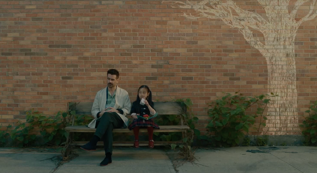 Jake et Mika sont assis sur un banc. Derrière eux se trouve un mur en brique sur le quel, à droite, est dessiné un arbre. Des plantes se trouvent entre le banc et le mur. Jake est à droite et à les jambes croisées. Mika est à gauche du banc et boit. De la nourriture se trouve sur ses genoux.