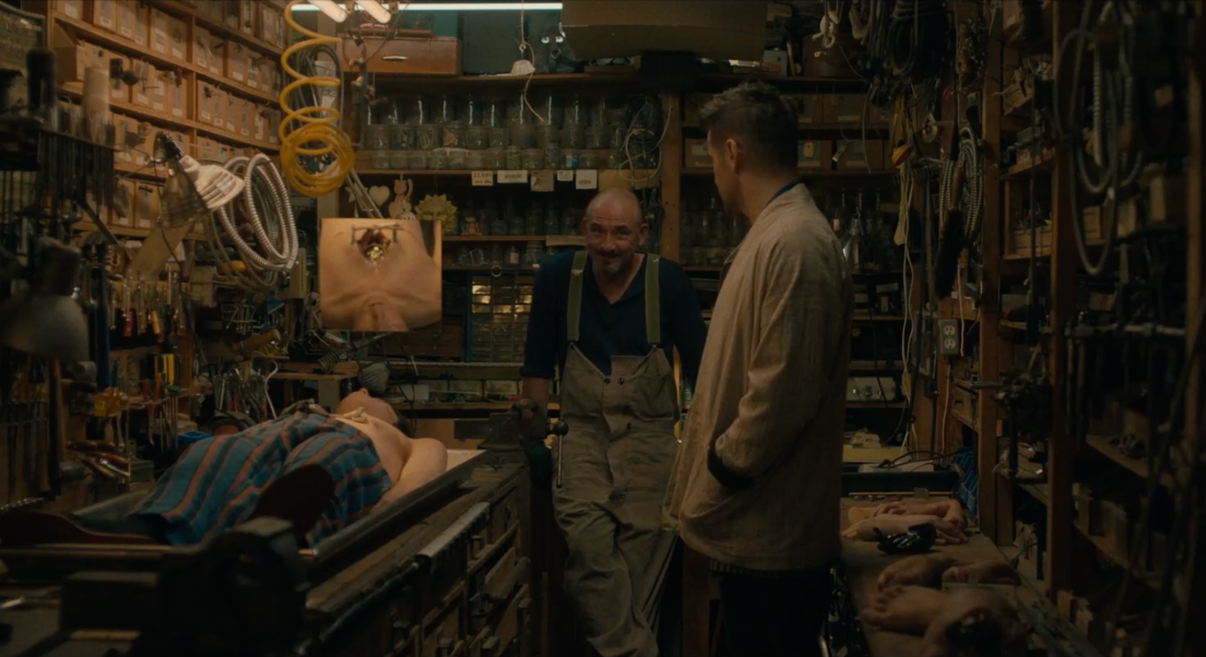 Jake et Yang se trouvent dans l'atelier de Ross. Yang est allongé sur une table. Ross est dans le fond de son atelier, discutant avec Jake. Ross nous fait face, Jake est de profil. Des outils sont présents toute la surface de tous les murs. Un miroir nous montre la partie du corps de Yang qui a été examinée.