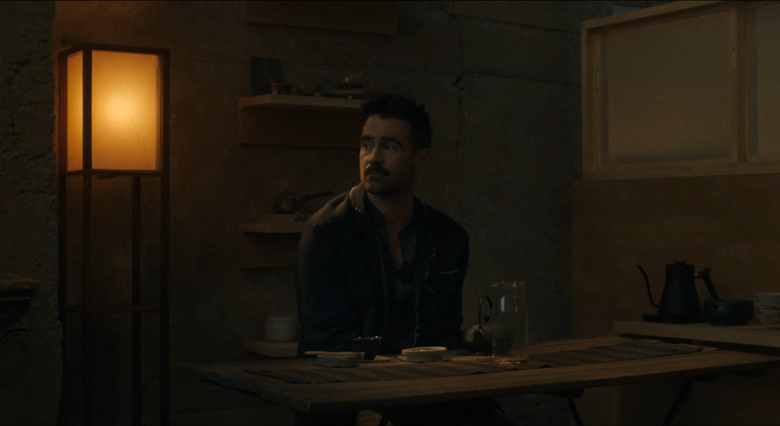 Jake est assis seul à un table et regarde vers la gauche. Devant lui se trouvent une théière, une bouteille d'eau et son thé. La pièce est dans la pénombre. Sur la gauche de Jake, une lampe est allumée. À sa droite, une deuxième théière est présente.