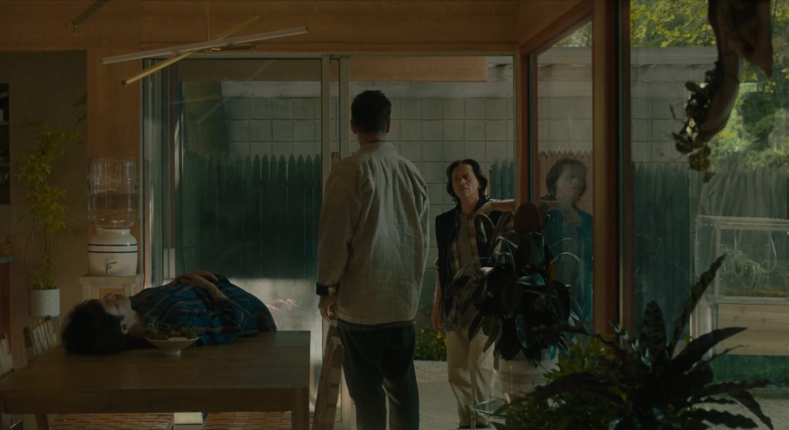 Jake discute avec son voisin George dans un pièce lumineuse. Il nous tourne le dos et George nous fait face, appuyé sur le cadrant de la porte. Yang est déposé sur la table, les mains coirsées sur son ventre. Des plantes se trouvent à droite de Jake.
