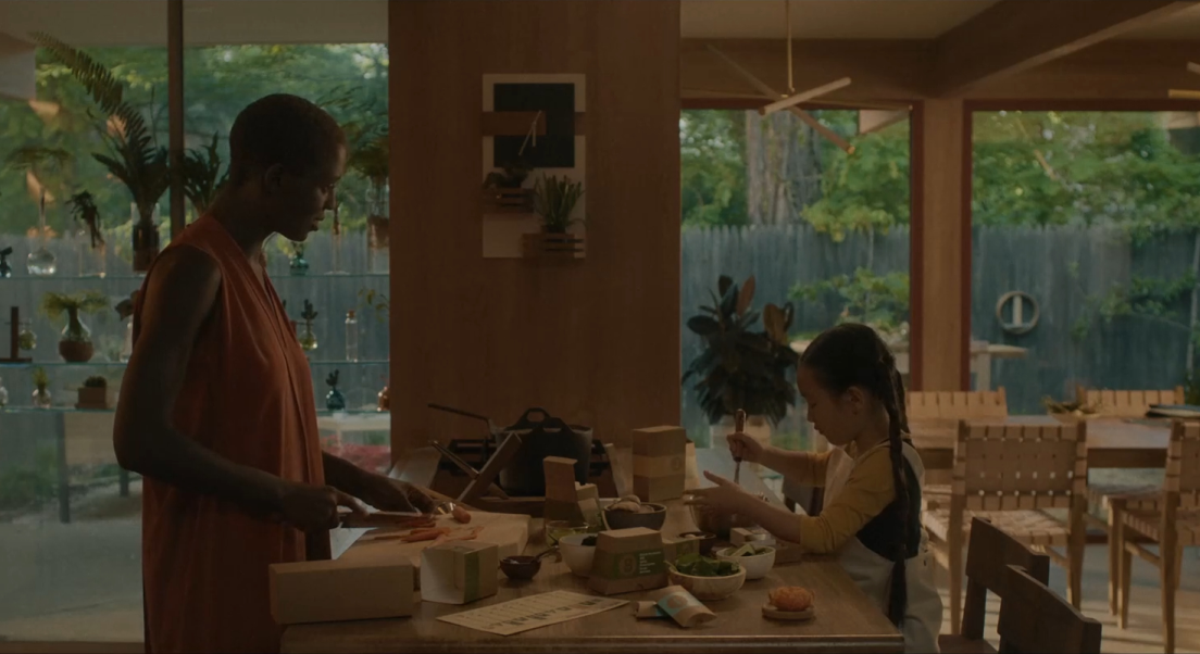 Kyra et Mika cuisine dans leur cuisine. Kyra coupe des carottes et Mika fouette dans un bol. Les ingrédients sont dispersés sur le plan de travail sur lequel elles cuisinent. Nous pouvons voir la table de la salle à manger en arrière plan, sur la droite.