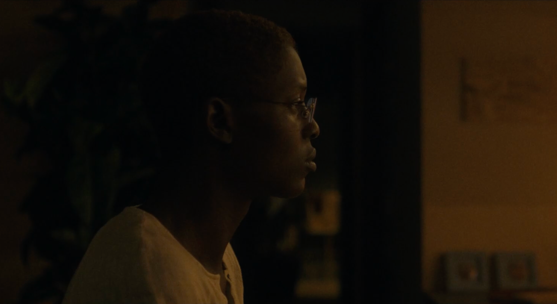 Kyra porte des lunettes et un chemiser blanc. Elle est situé de profil. La pièce dans laquelle elle se trouve est sombre.