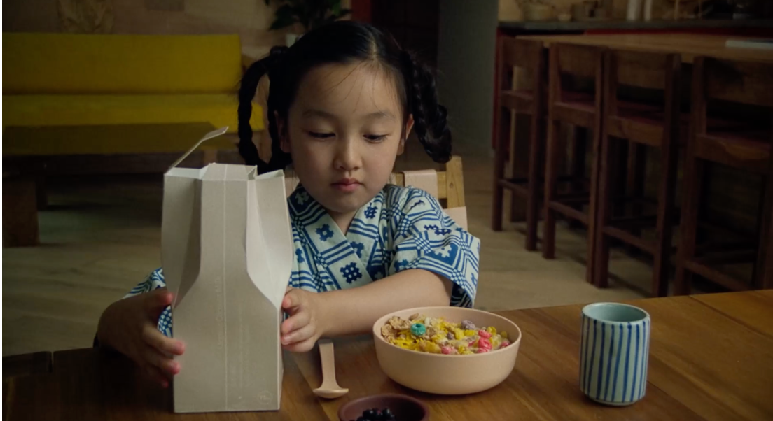 Mika est assise à une table et prend son petit déjouner. Devant elle se trouve un bol de céréal dans lequel elle va verser du lait. En arrière plan sur sa droite se trouvent une table avec des chaises. Sur sa gauche, se trouvent un canapé jaune et une table basse.