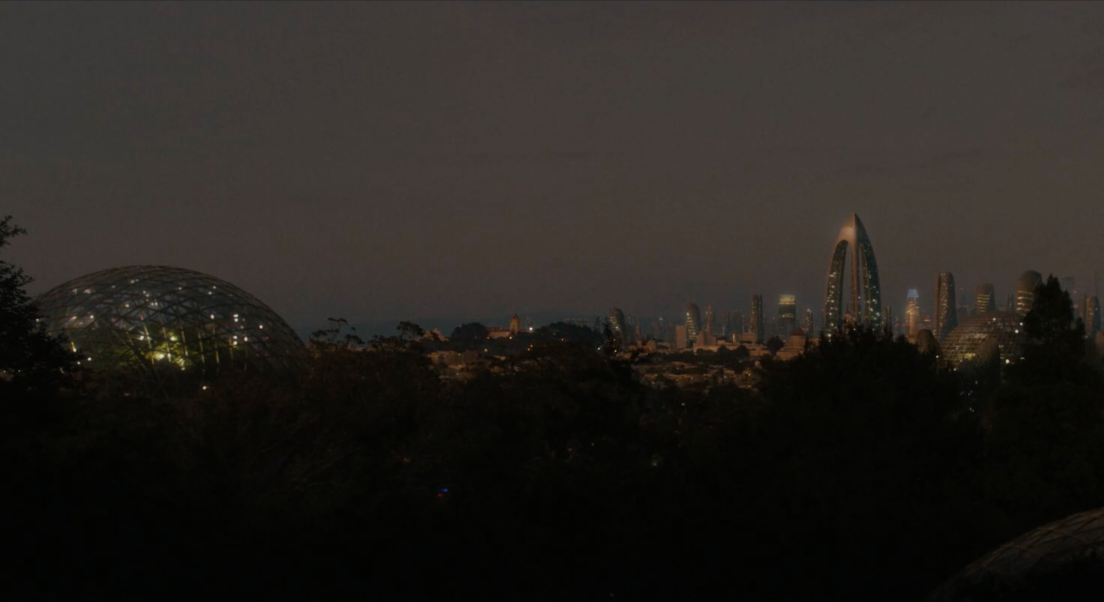 paysage futuriste d'une ville, à la tombée de la nuit. Sur la gauche, nous pouvons observer un bâtiments en dôme. Sur la droite, les bâtiments sont plus haut. Des arbres se trouvent en avant plan. La ville est illuminée.