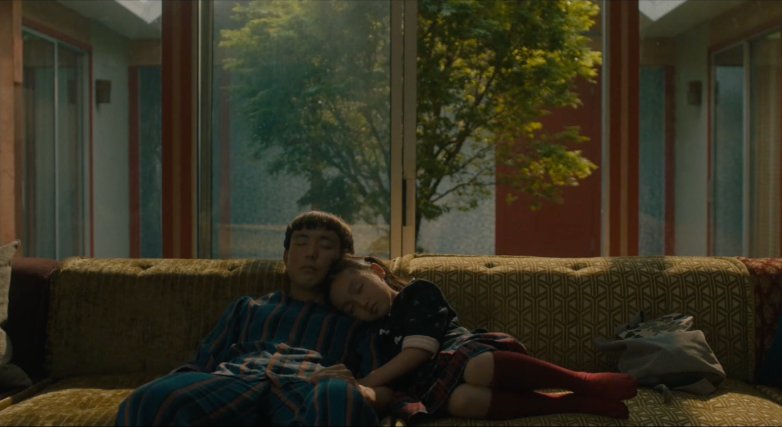 Mika et Yang dorment assis sur un canapé. Une baie vitrée se trouve derrière le fauteuil. À travers celle-ci, nous voyons un arbre dans une cour intérieure. Mika, à droite de l'image, a la tête posée sur l'épaule de Yang.