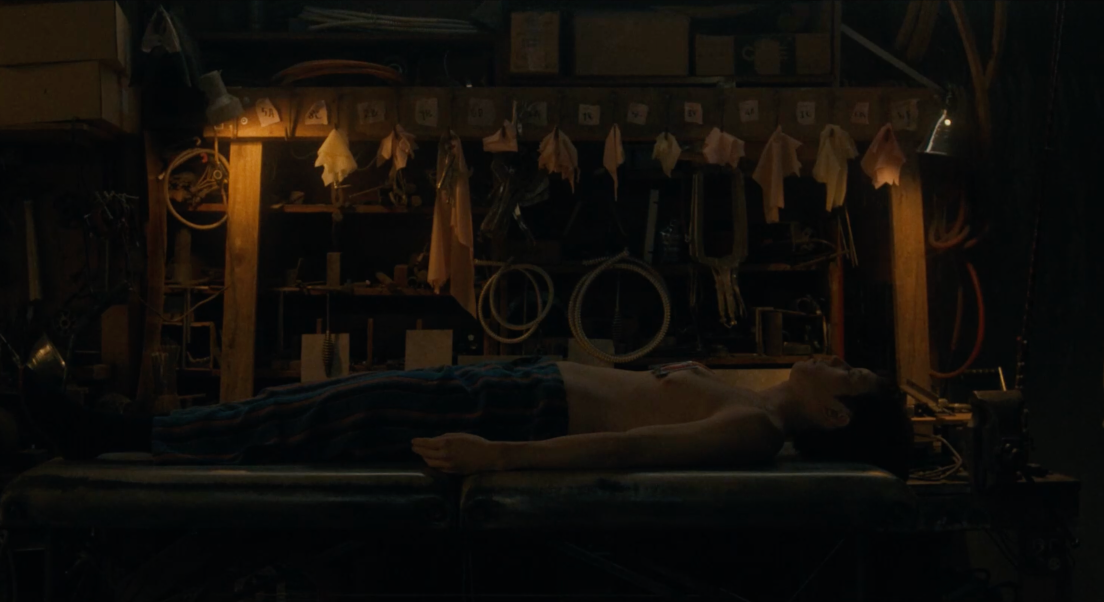 Yang est allongé sur une table. Derrière lui, des outils sont rangés sur une étagère en bois. La pièce est plongée dans la pénombre, avec deux lampes éclairant faiblement.