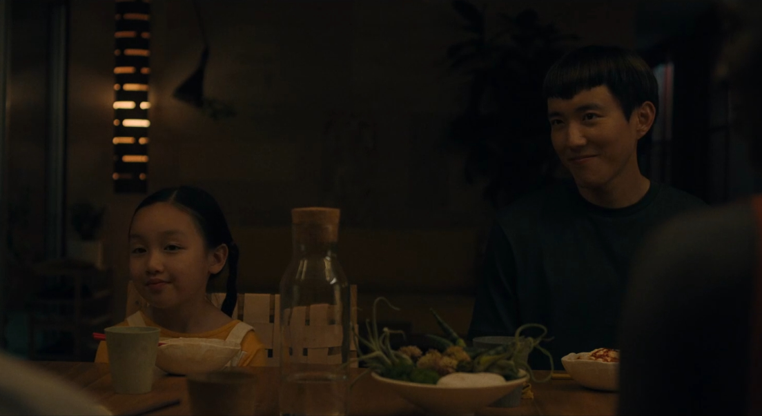 Mika et Yang sont assis à une table. Ils sont assis l'un à côté de l'autre, une assiette et une verre posés devant eux. Sur la table, une bouteille d'eau et de la décoration végétale sont présents. Yang et Mika nous font face.