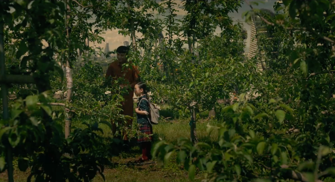Yang et Mika se tiennent debout dans un verger. Mika a un sac à dos et est de profil. Yang est de face et porte une salopette orange. Nous distinguons en arrière plan les bâtiments d'une ville.