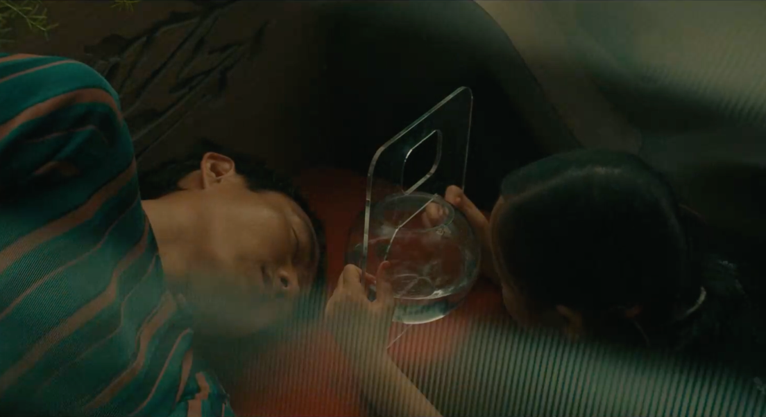 Yang est couché sur un siège. Mika tient devant sa tête un bocla avec un poisson. Elle regarde son poisson et Yang. Nous voyons la scène à travers une vitre.