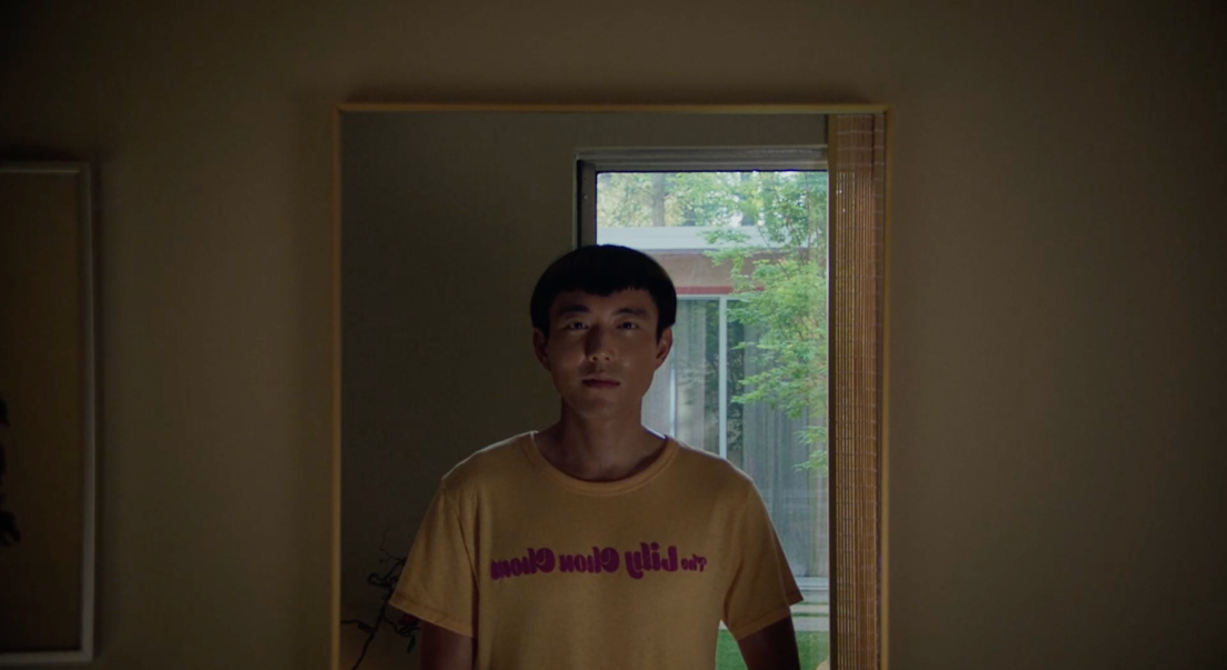 Yang se tient devant un mirroir. Derrière lui se trouve une baie vitrée donnant sur une cour intérieur. À l'intérieur de celle-ci, un arbre est présent.