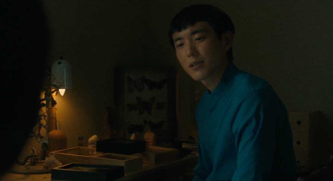 Yang se tient à côté d'un bureau sur lequel est exposé différentes boites avec des papillons. Une lampe posée sur ce bureau éclaire la pièce. Yang regarde vers le bas et porte un haut bleu.
