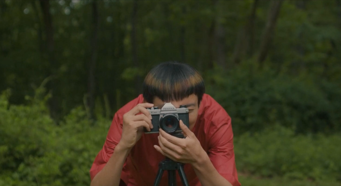 Yang est penché derrière sont appareil photo, posé sur un pied. Derrière lui se trouvent une forêt. Yang porte une tenue rouge.
