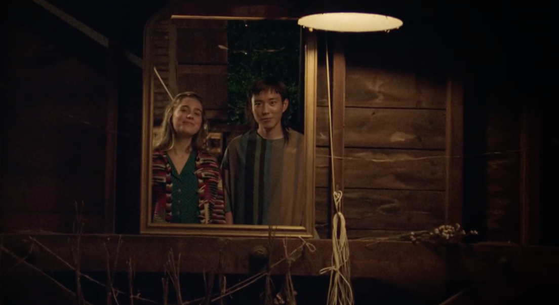 Yang avec des cheveux longs et une jeune fille se regardent dans un mirroir. Le miroir est déposé sur une poutre d'un mur en bois. Yang et la jeune fille se trouvent à l'intérieur d'une maison en bois.