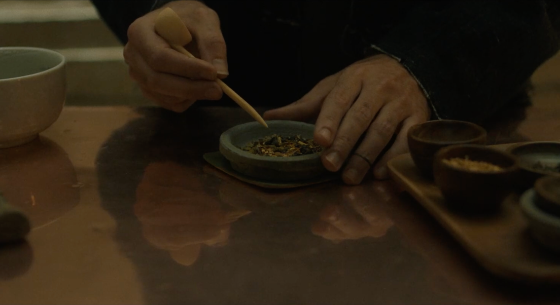 Focus sur les mains de Jake qui prépare son thé. Il tient une pique en bois et écrase les ingédients du thé. D'autres composants sont dans des petits bols, sur un plateau situé sur sa droite. Un bol blanc se trouve sur sa gauche.