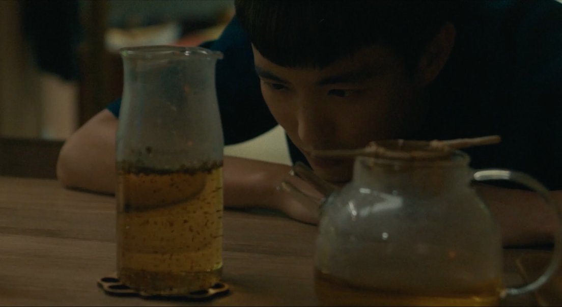 Yang regarde une bouteille en verre contenant du thé. Il a sa tête appuyée sur ses bras qui se trouvent sur la table. À côté de la bouteille ets posée une théière.